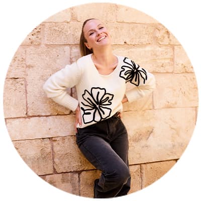 Frau in weißem Pullover mit schwarzen Blumendruck lehnt an einer Steinwand, Hände in den Hüften