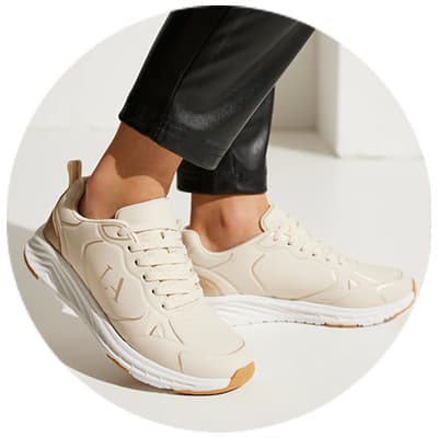 Beige Sneaker mit weißer Sohle, getragen von Person mit schwarzer Hose, Detailansicht der Schuhe auf weißem Hintergrund