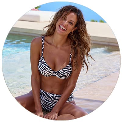 Frau im Zebra-Bikini sitzt am Poolrand, schaut in die Kamera und lächelt, im Hintergrund Wasser und blauer Himmel