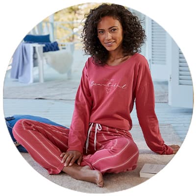 Frau in rosa Pyjama mit Schriftzug „Beautiful“ sitzt barfuß entspannt auf einer Veranda