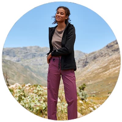 Frau in Outdoorhose und Jacke steht lächelnd in bergiger Landschaft mit weitem Tal im Hintergrund