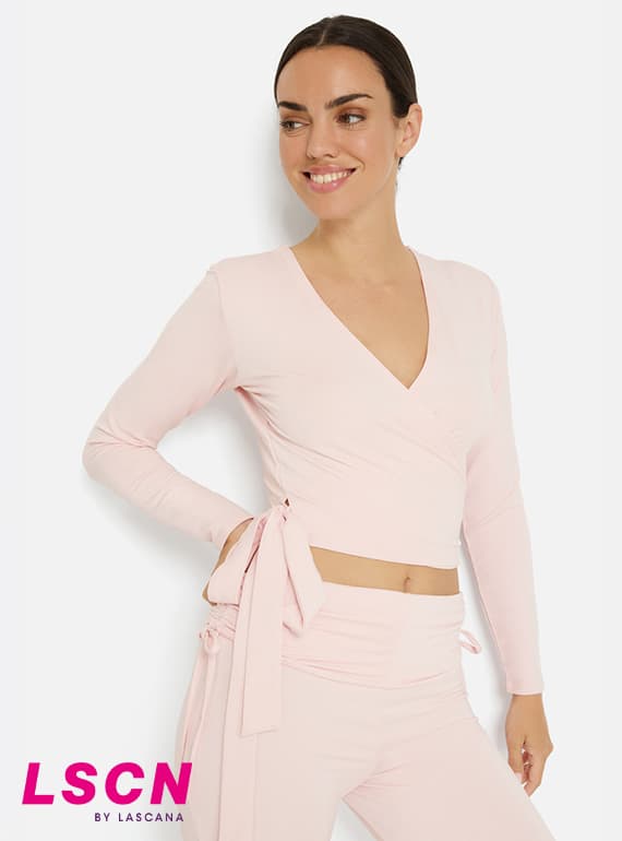 LSCN by Lascana – Frau im rosa Wickel-Longsleeve und passender Hose steht seitlich und blickt lächelnd nach links