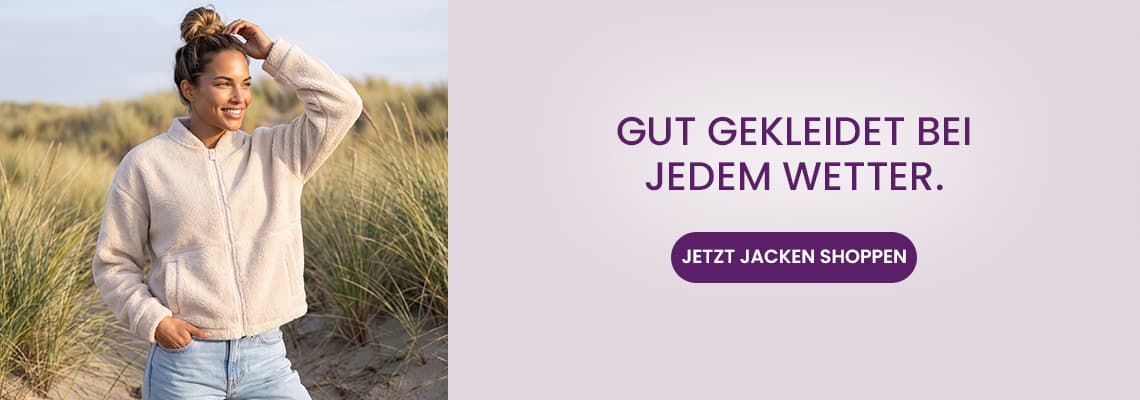 Gut gekleidet bei jedem Wetter – Frau in heller Jacke steht am Strandgras und schaut lächelnd zur Seite