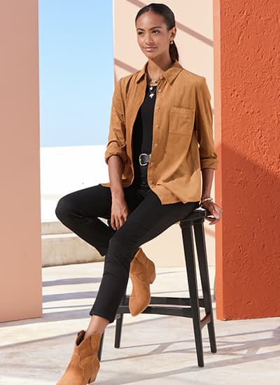 Frau in schwarzer Hose und schwarzem Top mit offener brauner Bluse sitzt auf Hocker vor orangefarbener Wand