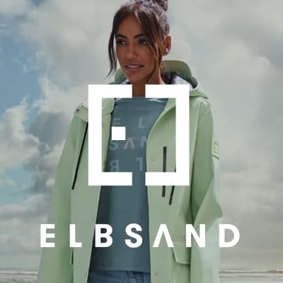 ELBSAND – Frau trägt mintgrüne Jacke über blauem Shirt mit ELBSAND-Aufdruck vor bewölktem Himmel.