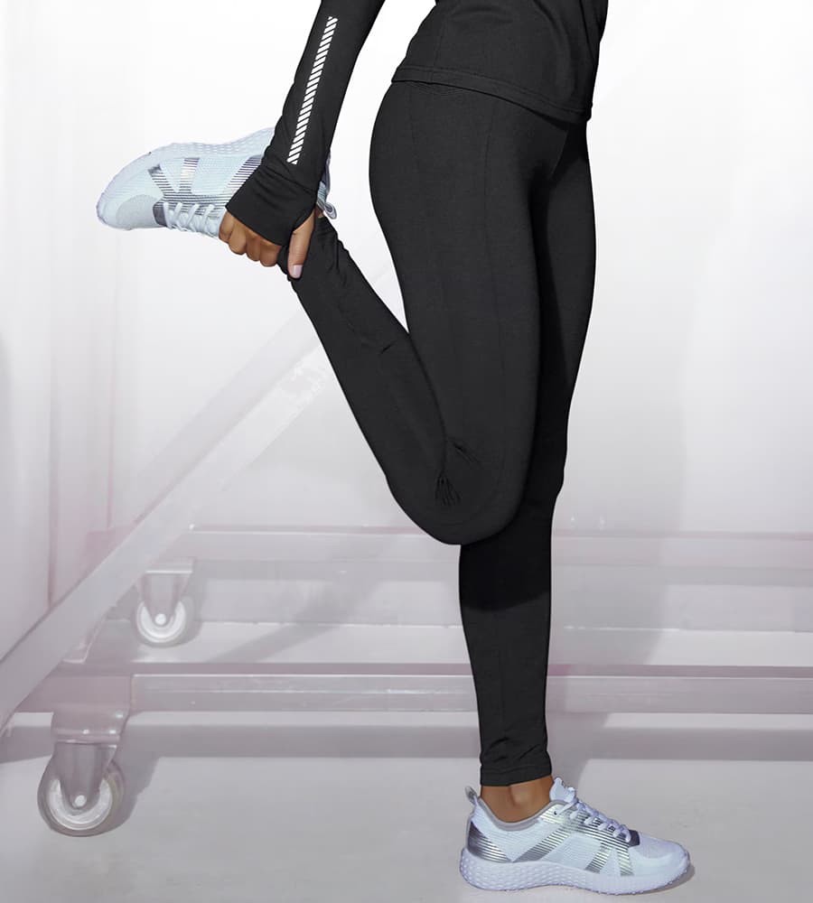 Frau in schwarzer Sportkleidung zieht ein Bein in Dehnübung an, trägt weiße Turnschuhe.