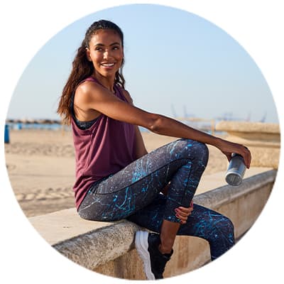 Frau in Sportkleidung sitzt auf Mauer am Strand, hält eine Trinkflasche in der Hand.
