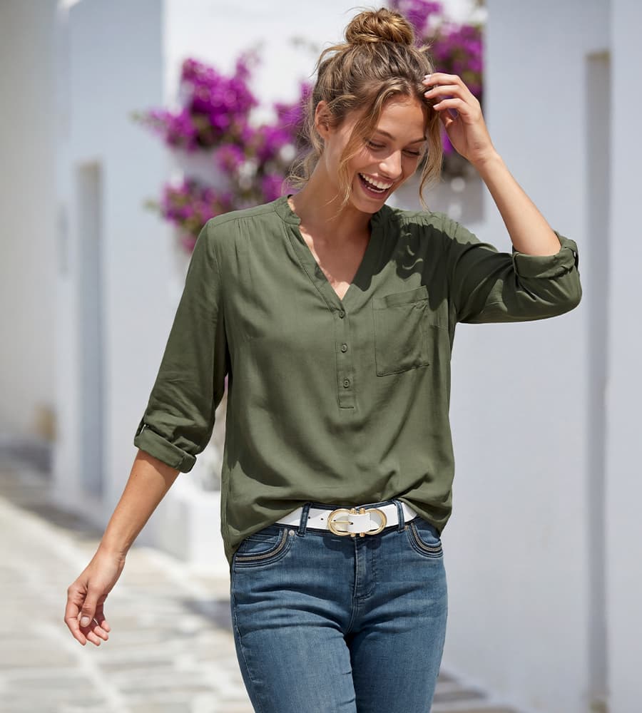 Frau in olivgrüner Bluse und Jeans mit weißem Gürtel geht durch helle Gasse mit lila Blumen im Hintergrund