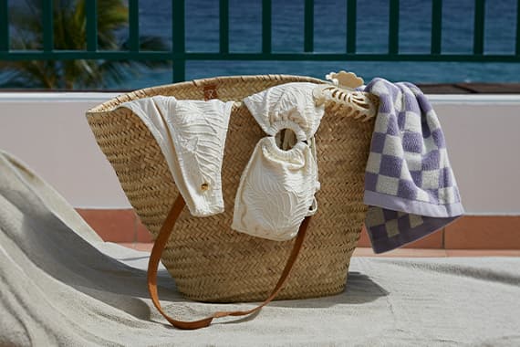 Geflochtene Strandtasche mit weißem Bikini und kariertem Handtuch, im Hintergrund Meer und grüne Balkonstreben